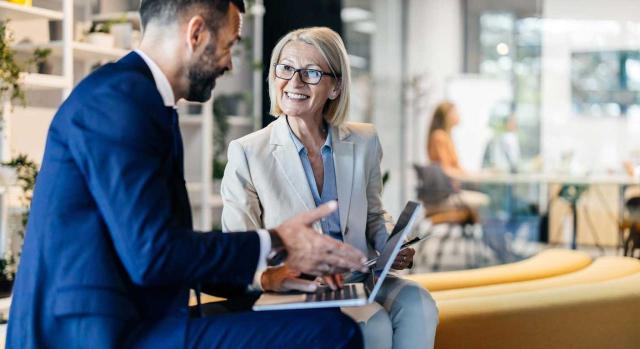 Aportaciones no dinerarias en las operaciones societarias: contabilización y tratamiento fiscal Aportaciones no dinerarias en las operaciones societarias: valoración y tratamiento contable. Imagen de una pareja de empresarios hablando muy sonrientes en su puesto de trabajo